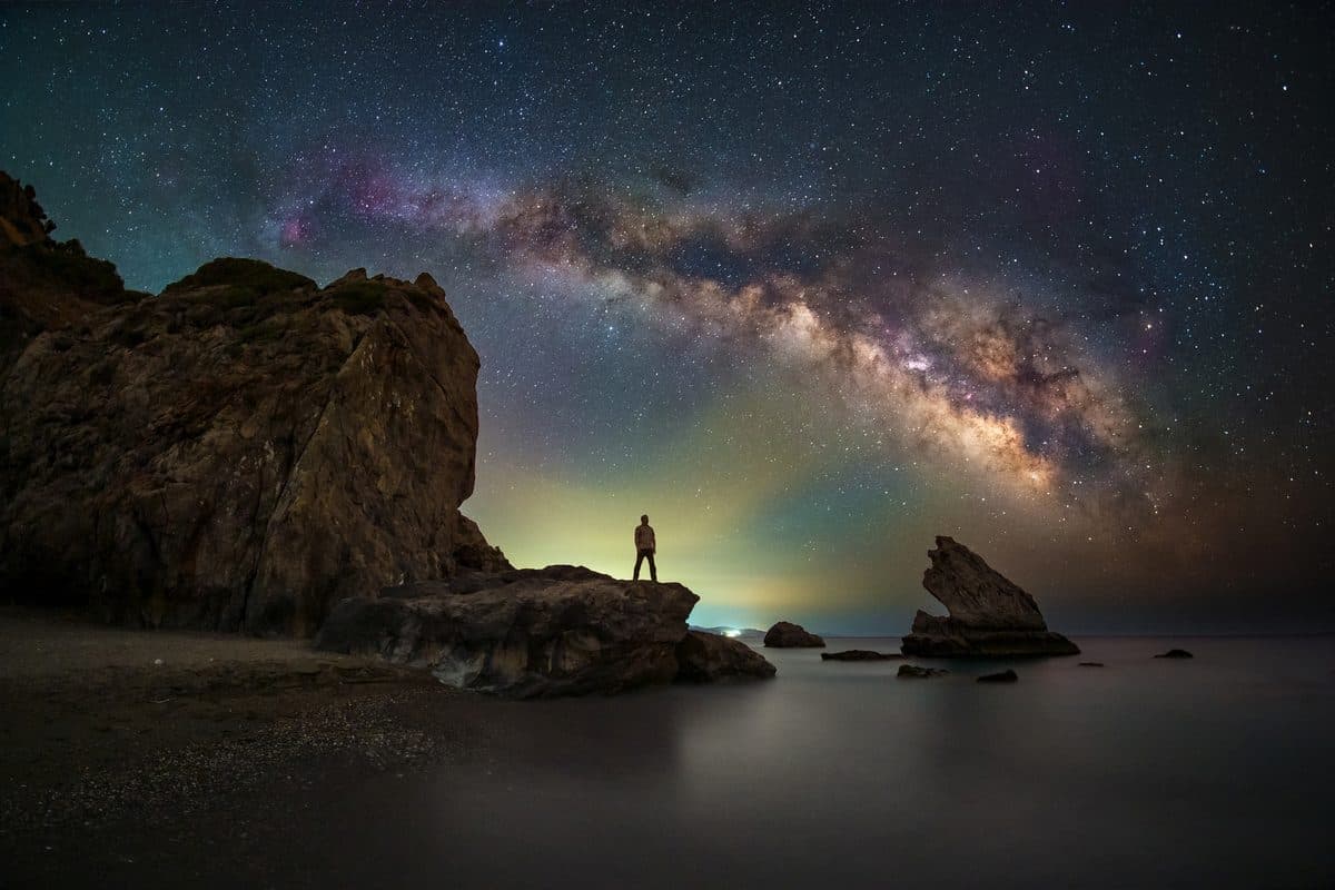 Silhouetted figure on coastal rocks under the Milky Way arch by Evgeni Tcherkasski